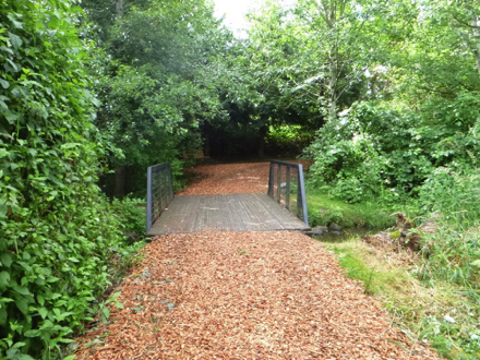 Short wooden foot bridge with railings crosses small creek - bark chip trail leads into a forest